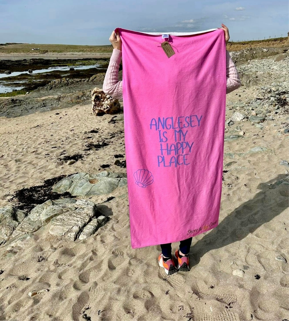 alt=“Pink microfibre beach towel with the slogan ‘Anglesey is my happy place’ in bold white text – lightweight, quick-drying, and perfect for coastal days with Shore & Coast”
