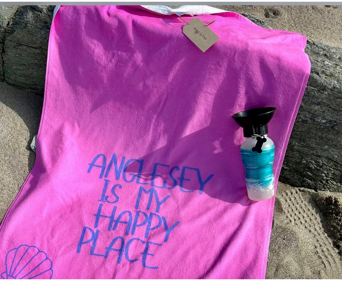 alt=“Pink microfibre beach towel with the slogan ‘Anglesey is my happy place’ in bold white text – lightweight, quick-drying, and perfect for coastal days with Shore & Coast”