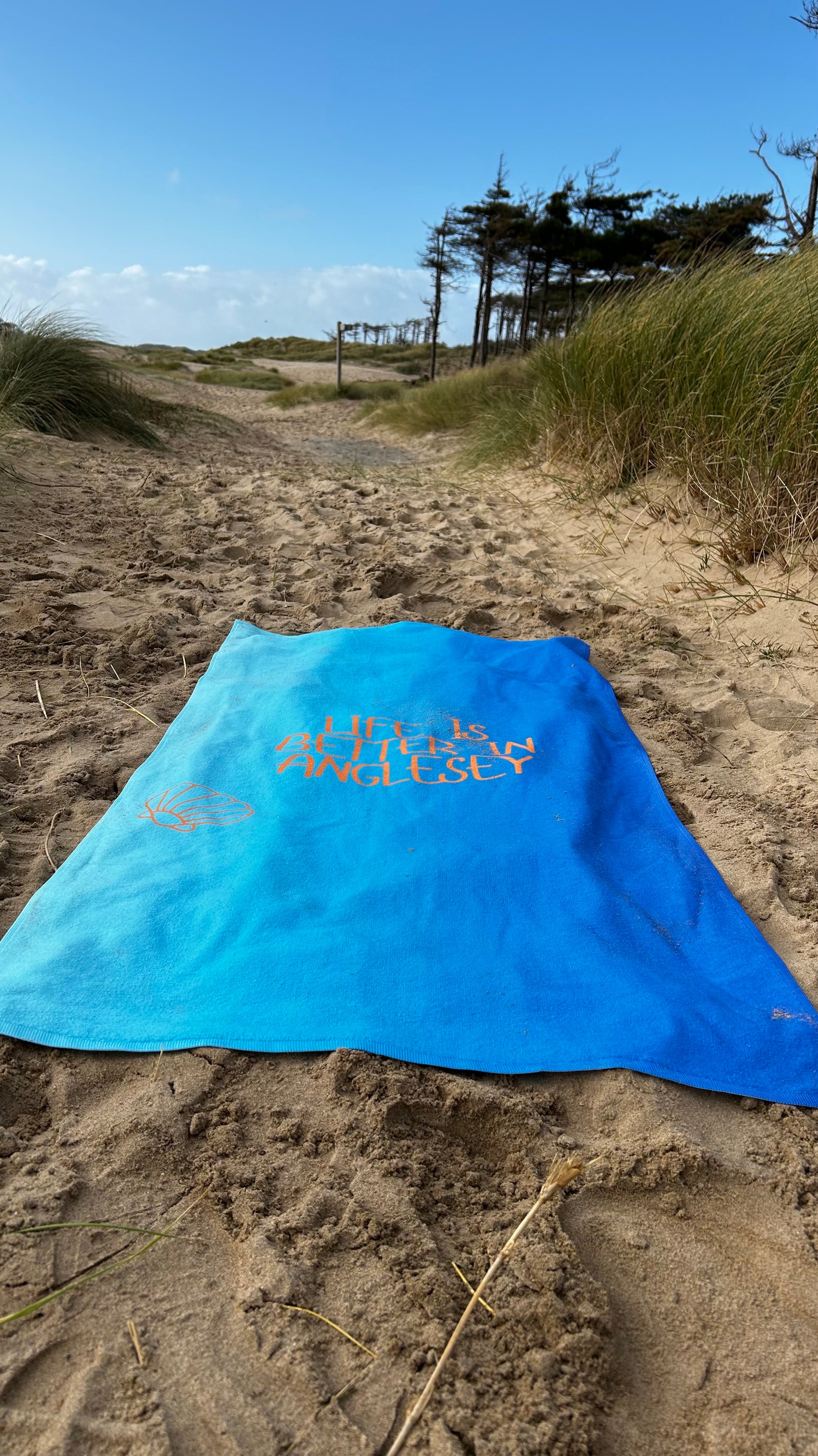 alt=“Vibrant blue microfibre beach towel with the slogan ‘Life is better in Anglesey’ in bold white text – lightweight, quick-drying, and perfect for beach adventures by Shore & Coast”