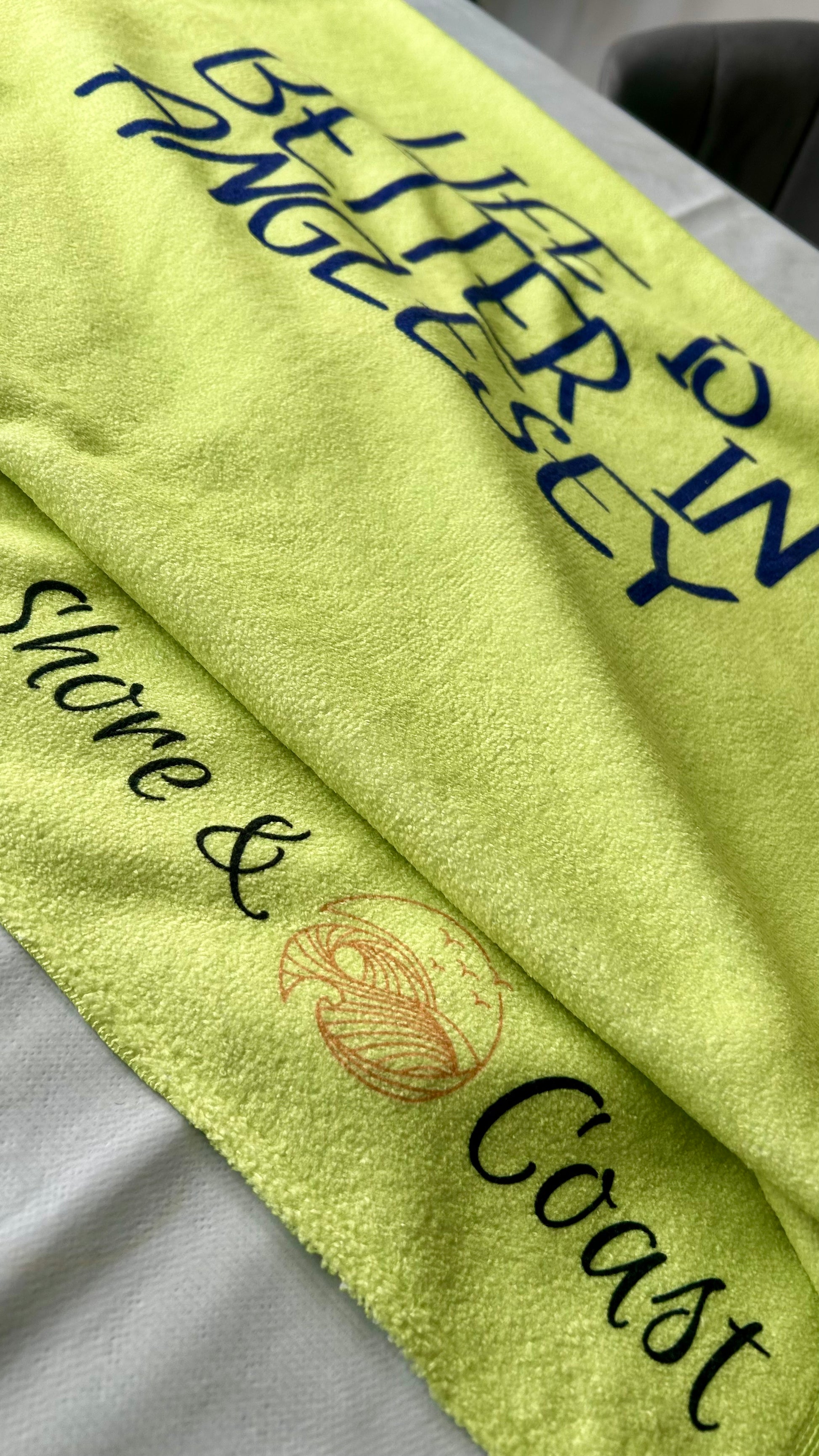 alt=“Lime green microfibre beach towel with cotton loops and the slogan ‘Life is better in Anglesey’ in bold yellow text – ultra-absorbent, quick-drying, and perfect for seaside adventures”