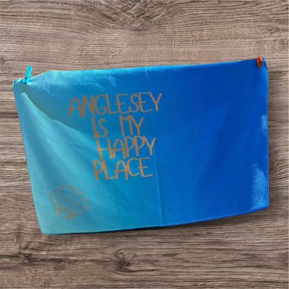 alt=“Quick-drying and absorbent microfibre tea towel with ‘Anglesey is my happy place’ slogan – ideal for beach lovers and coastal kitchens”