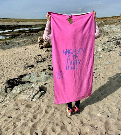 alt=“Pink microfibre beach towel with the slogan ‘Anglesey is my happy place’ in bold white text – lightweight, quick-drying, and perfect for coastal days with Shore & Coast”